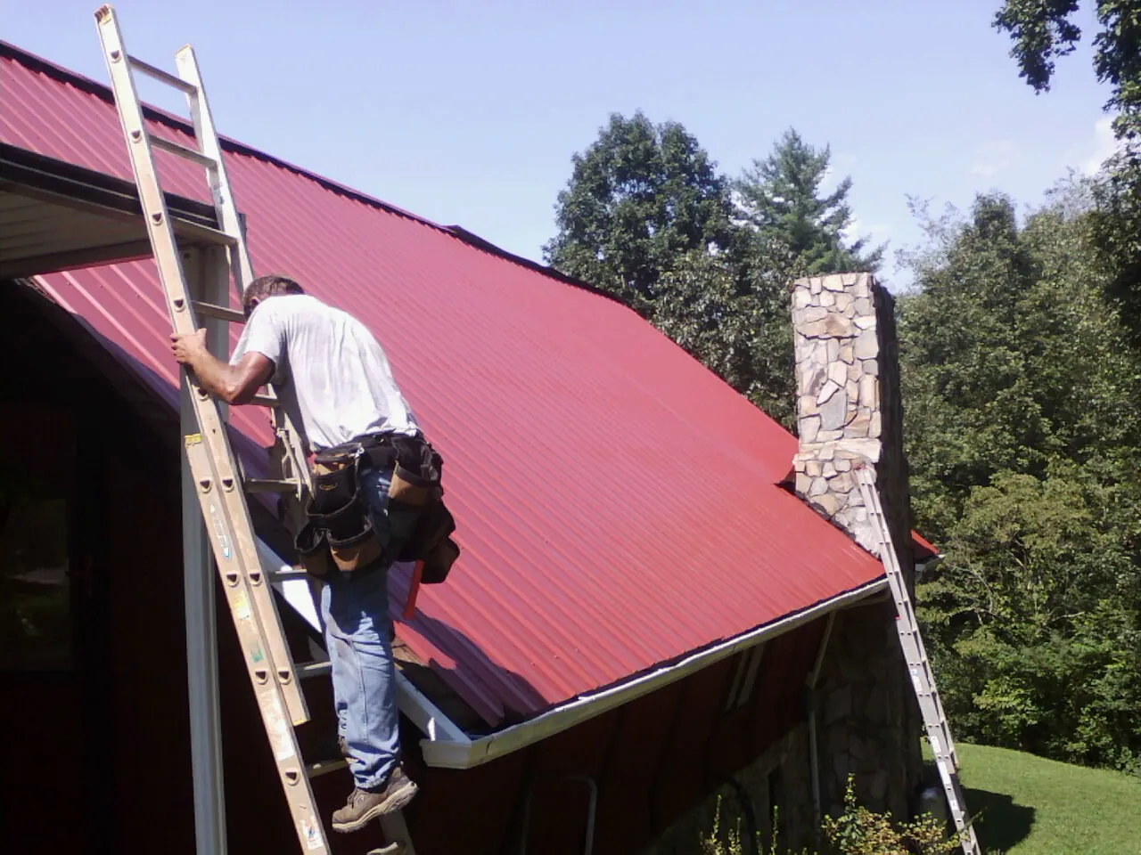 Roofer on a ladder beside a red metal roof and stone chimney — Soffit & Fascia Repair by Roofing Brothers in Fish Haven